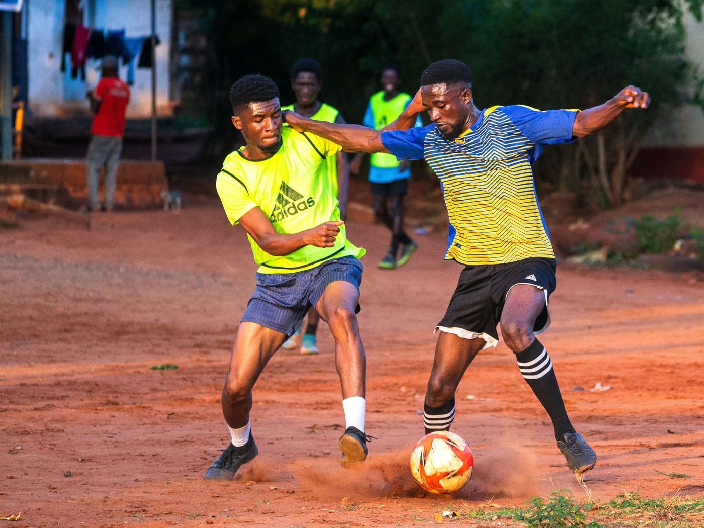 A grassroots five-a-side game on a red-dirt pitch in West Africa, two players in bibs battling for the ball